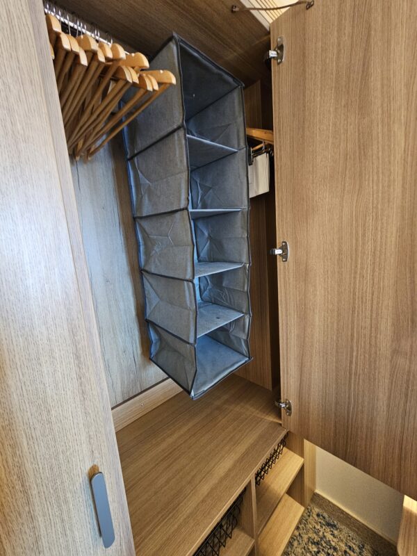 Open cruise cabin closet with a fabric hanging shelf organizer suspended beside a row of wooden hangers. The organizer hangs above wooden shelving with black wire baskets. This photo illustrates a practical cruise cabin hack that maximizes closet space and keeps small staterooms organized and functional.