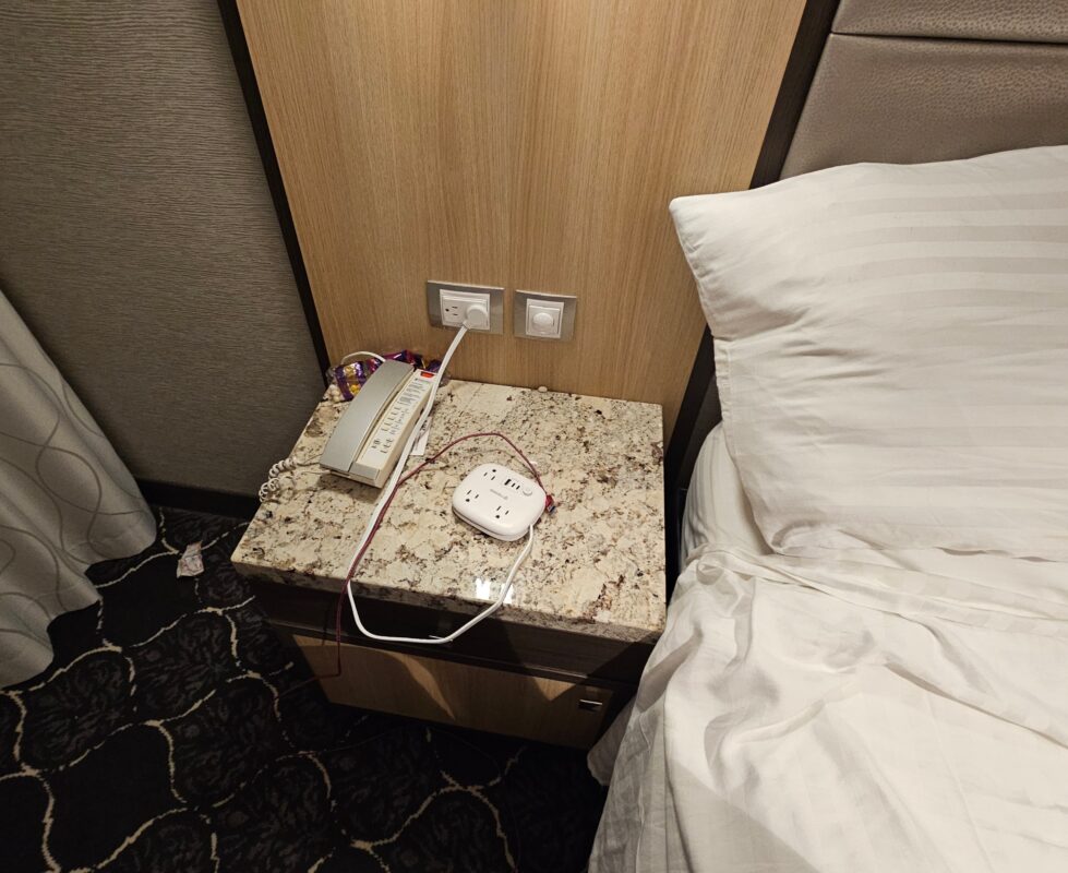 Close-up of a cruise cabin nightstand with a granite top, showing a wall outlet with a compact white outlet expander plugged in. A charging cable is connected, and a cruise phone sits nearby. This image illustrates a tech-friendly cruise cabin hack that helps travelers manage limited power outlets for multiple devices.