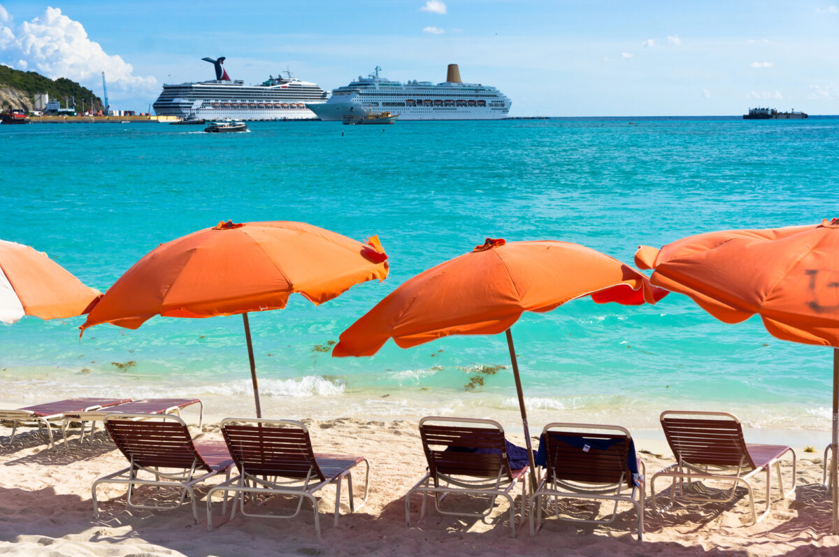 A row of orange beach umbrellas on Great Bay Beach, one of the St. Maarten beaches near cruise port in Philipsburg, St. Maarten, facing calm blue water and docked cruise ships in the background.