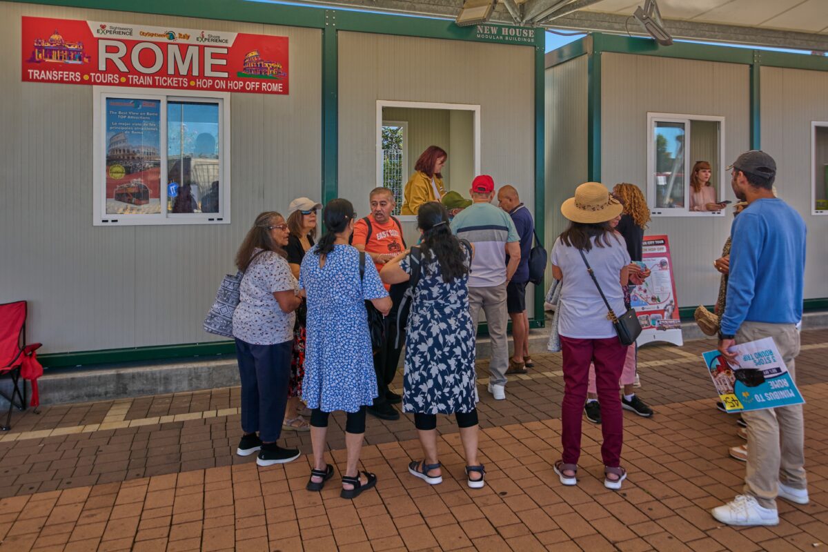 Travelers stand in line outside a modular building with signage for Rome tours and transfers, emphasizing the need for clear plans at foreign cruise terminals.