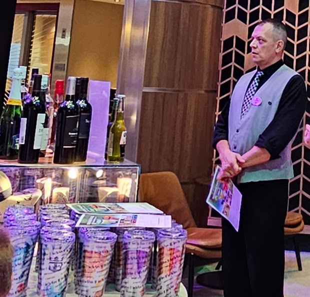 Staff member presenting drink package options near a table with branded souvenir cups and bottles of wine and beer.
