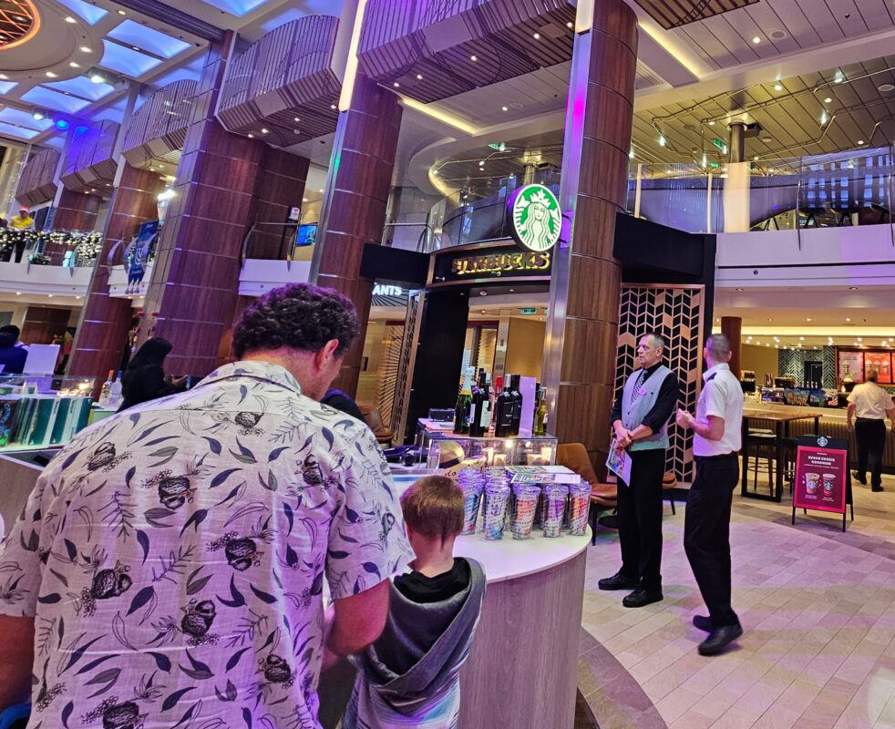 Is a Royal Caribbean Drink Package Worth It | Royal Caribbean crew members standing near a drink package sales station by the Starbucks café, greeting guests on embarkation day—a time when prices are typically higher.