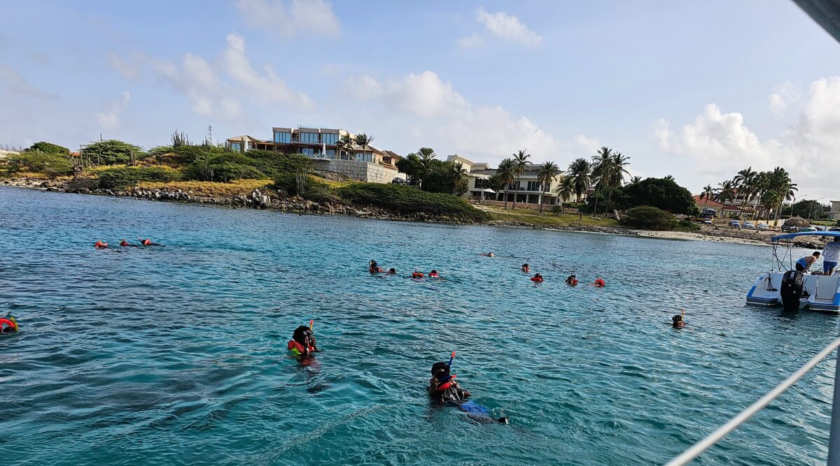 Snorkelers floating in turquoise water off the rocky coast of Aruba, wearing life vests and snorkel gear near a catamaran boat.