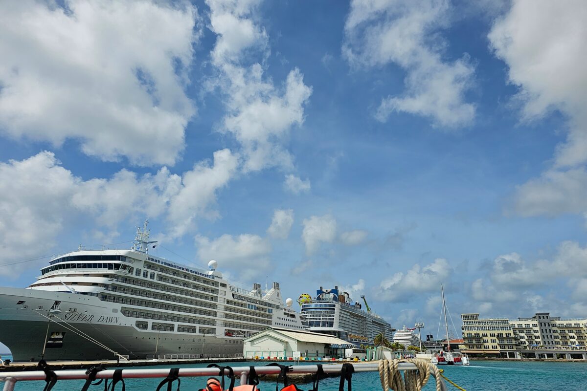 Panoramic view of several large cruise ships at the Aruba cruise terminal, with downtown buildings and catamarans nearby.