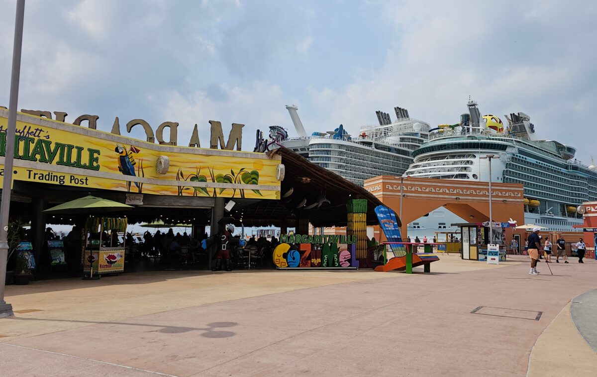Cruise passengers at the Cozumel pier area with a brightly colored Margaritaville bar in front and docked ships in the distance.