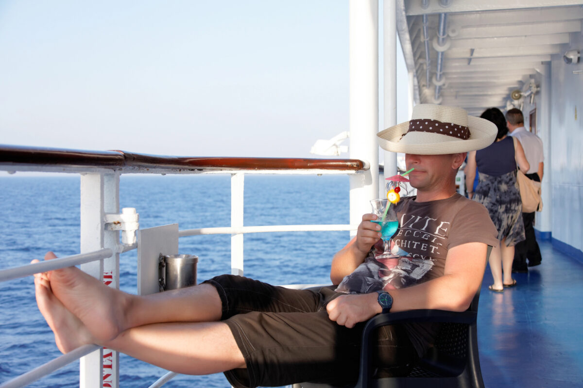 Person relaxing by the ship railing with a bright blue drink, festive fruit garnish, and straw hat obscuring their face.