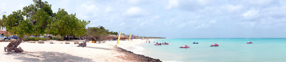 Panoramic view of Eagle Beach in Aruba with leaning divi-divi trees, jet skis on the water, and people walking along the shoreline.