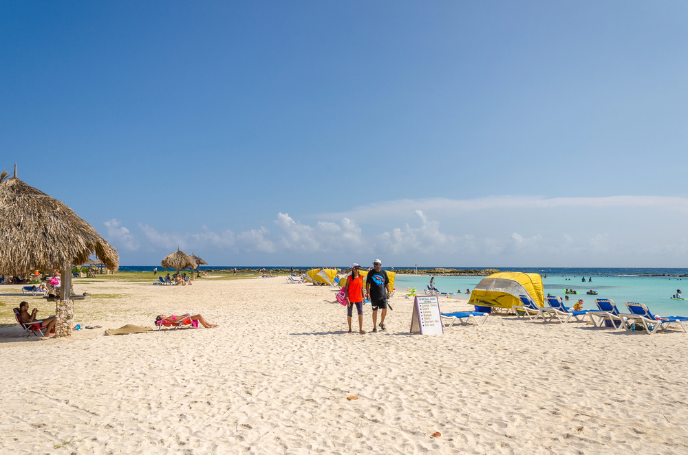 Shallow, sheltered waters of Baby Beach in Aruba with beachgoers wading and relaxing near colorful shade tents and palm-covered huts.