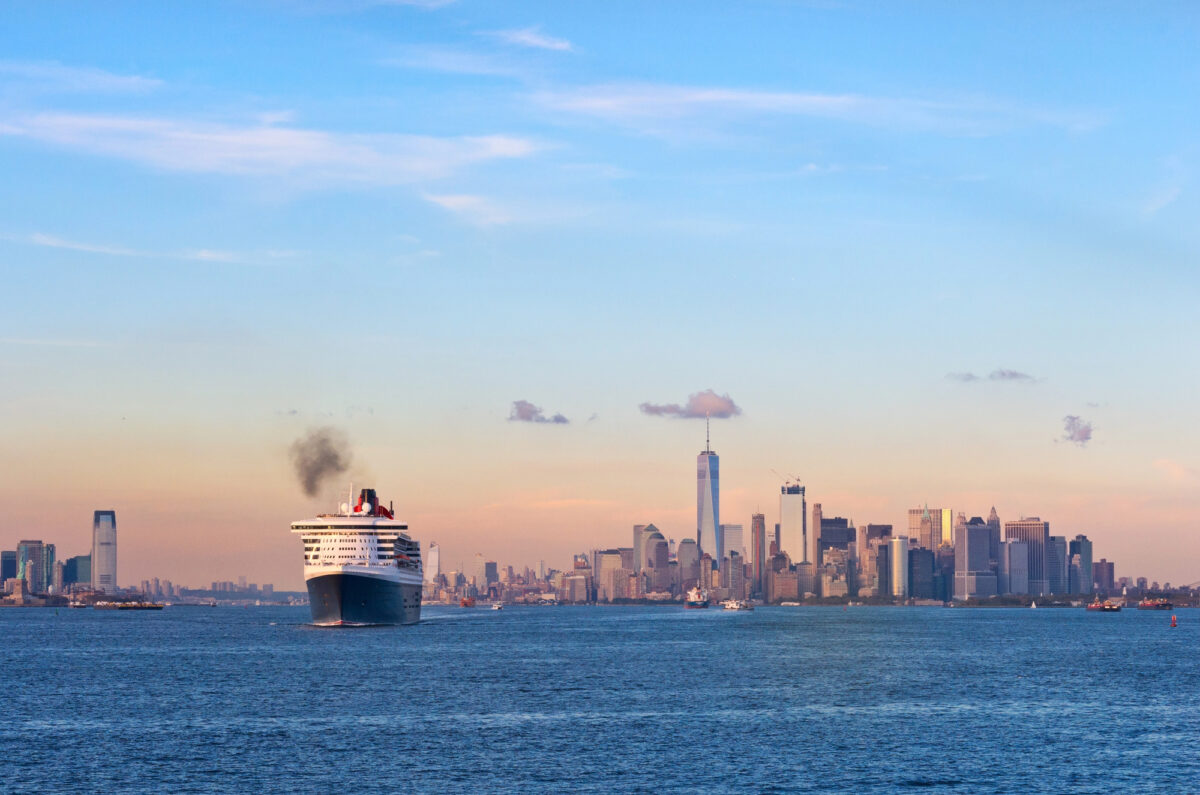 Cunard’s Queen Mary 2 sailing from New York Harbor on a transatlantic voyage, with the Lower Manhattan skyline and One World Trade Center in the background.