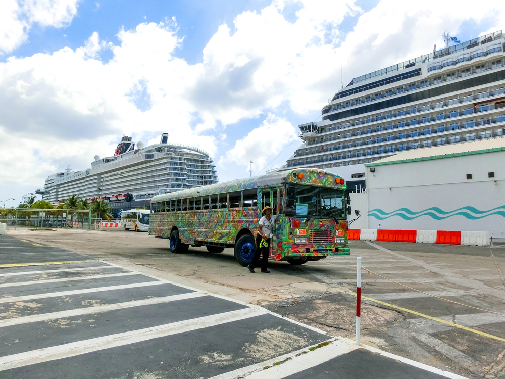 Vibrantly decorated bus in front of large docked cruise ships in Oranjestad, Aruba, on a partly cloudy day.