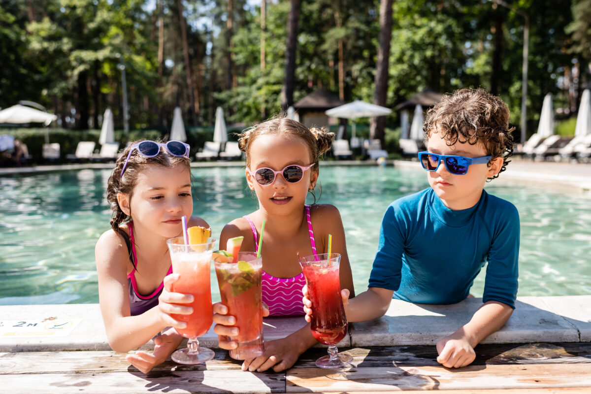 Kids enjoying mocktails with fruit garnishes at a resort-style pool, wearing bright swimsuits and summer gear.