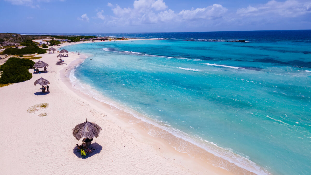 Aerial view of Baby Beach in Aruba with turquoise water, white sand, and scattered thatched palapas providing shade to beachgoers.