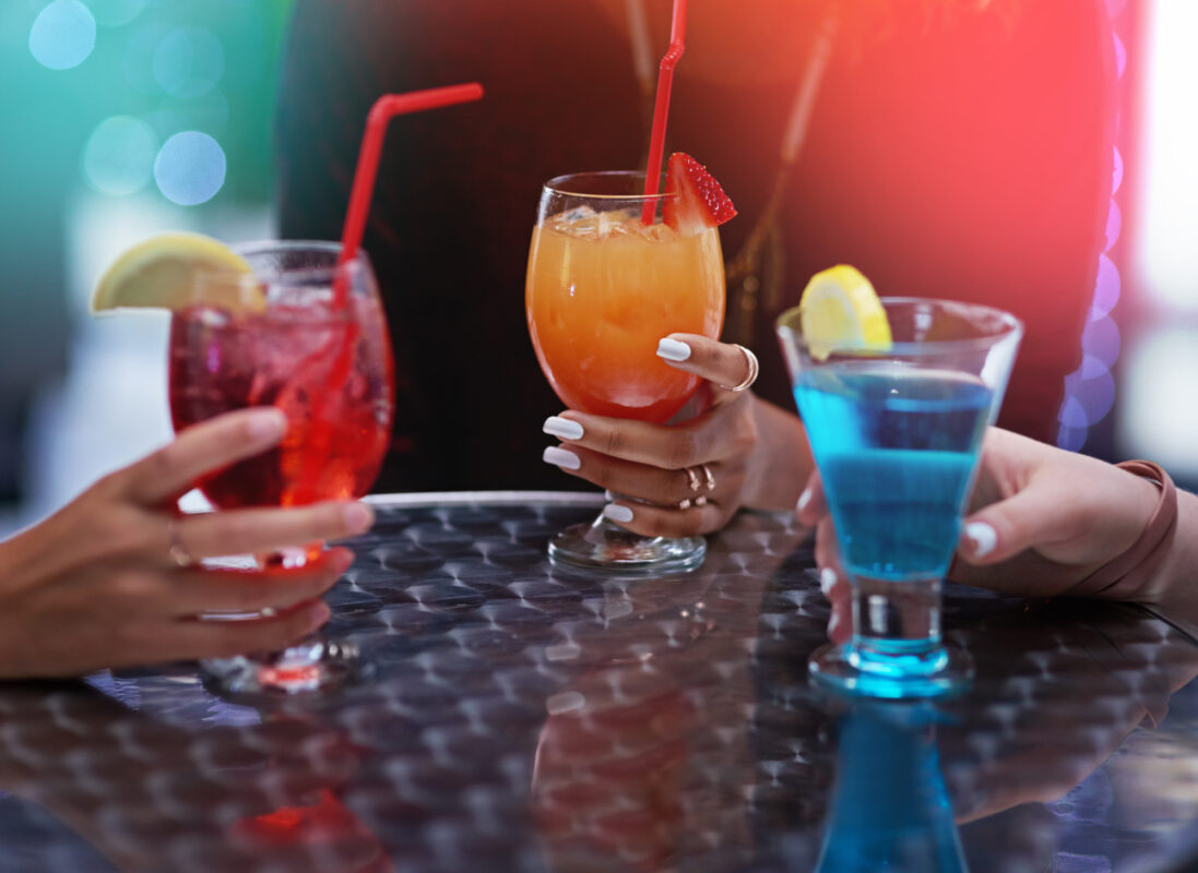 Close-up of hands holding mixed drinks with fruit garnishes and straws on a cruise-style metal table enjoying cruise drink packages.