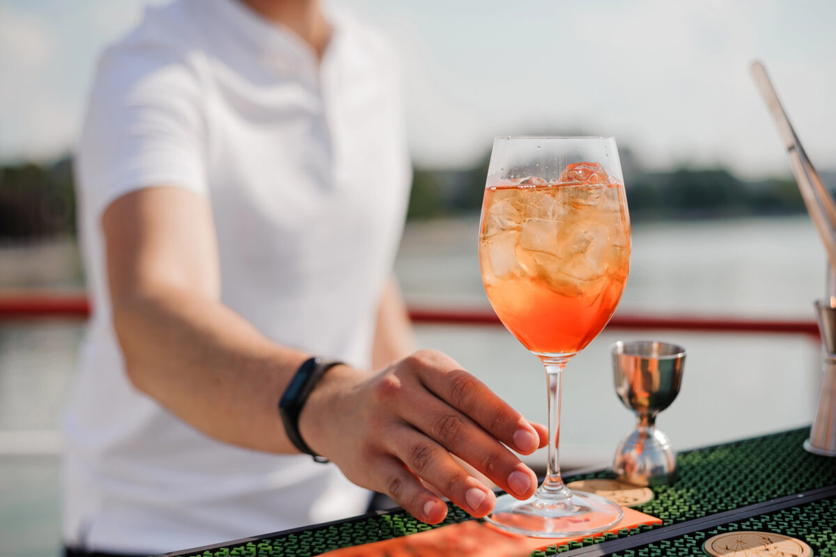 A close-up of a bartender handing a guest an orange-hued iced cocktail in a wine glass, set against a blurred waterfront background.