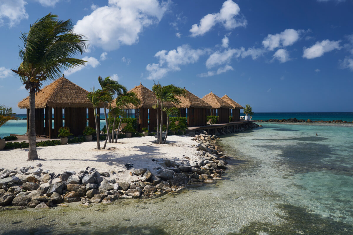 Palm-fringed private cabanas on a sandy point with rocky shoreline and clear water on Aruba’s Renaissance Island.