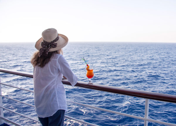 Woman in a sun hat standing on a cruise ship balcony with a tropical cocktail on the railing, gazing out at the open sea.