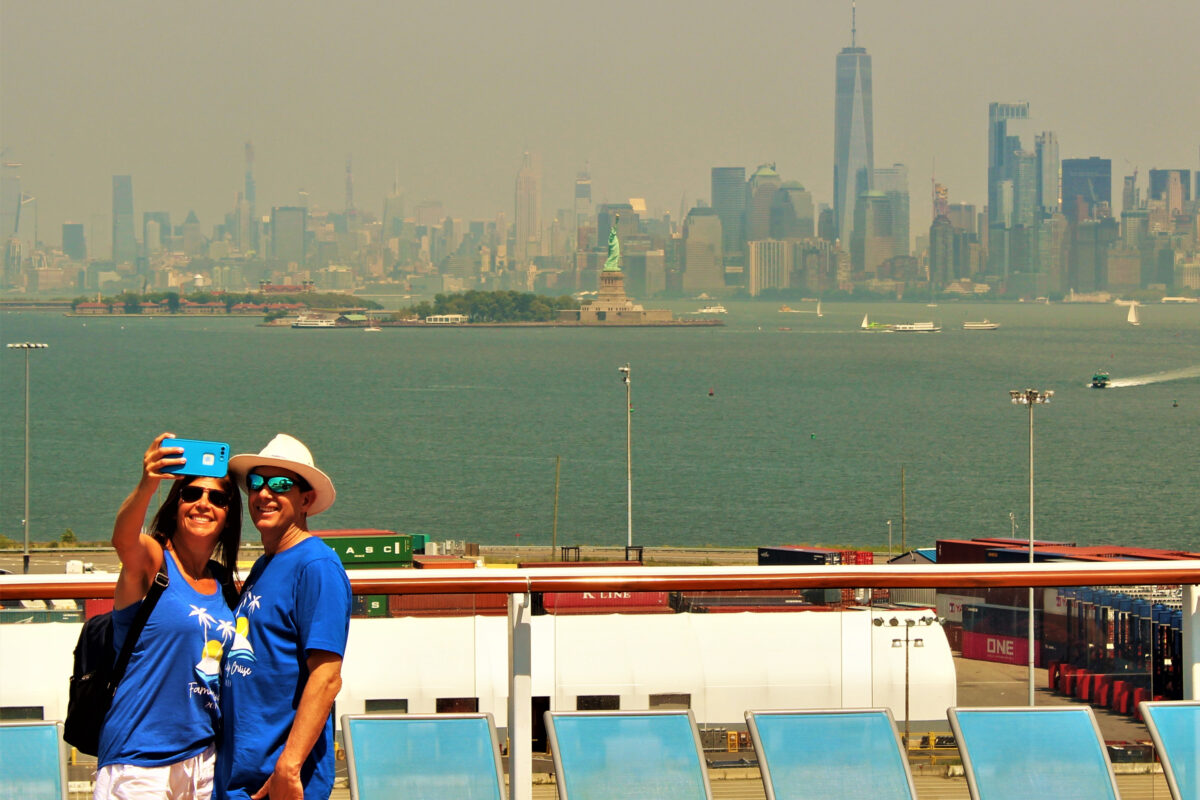 Cruise passengers take a selfie on deck with the Statue of Liberty and New York City skyline in the background, departing from Cape Liberty in New Jersey.