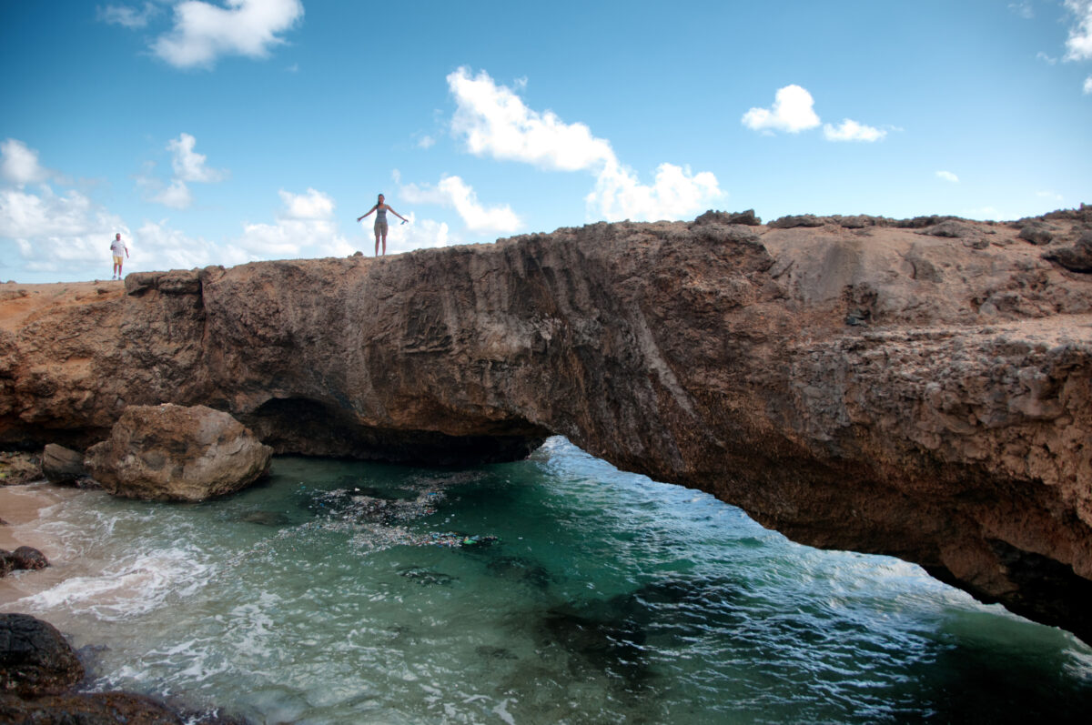 Tourists standing on a small natural limestone bridge over turquoise water on the rocky northern coast of Aruba.
