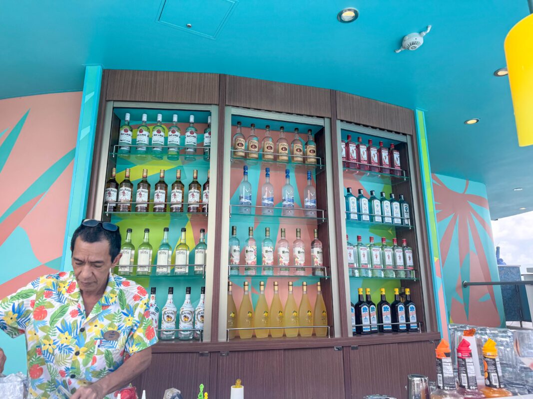 A bartender prepares drinks at a Royal Caribbean Lime & Coconut bar, with shelves of spirits and a colorful tropical design in the background.