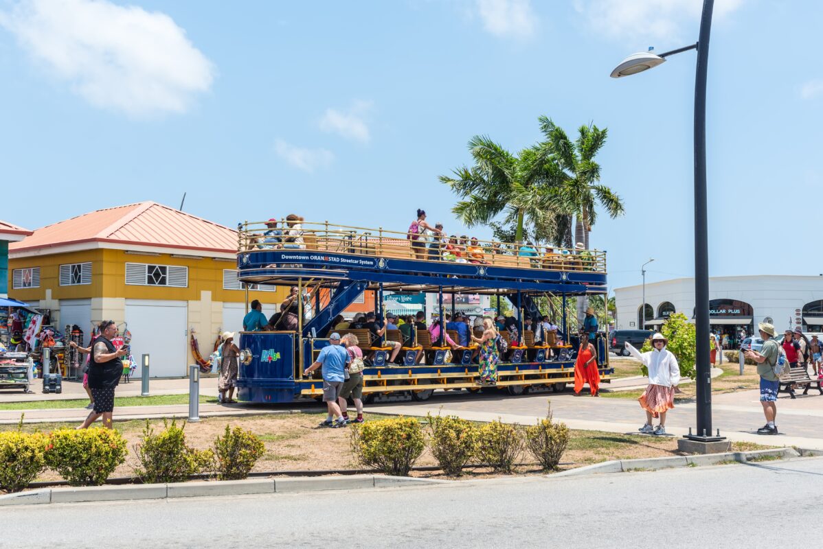 Tourists riding an open-air, double-decker streetcar in downtown Oranjestad, Aruba, with shops and palm trees in the background. One of the free things to do in Aruba.