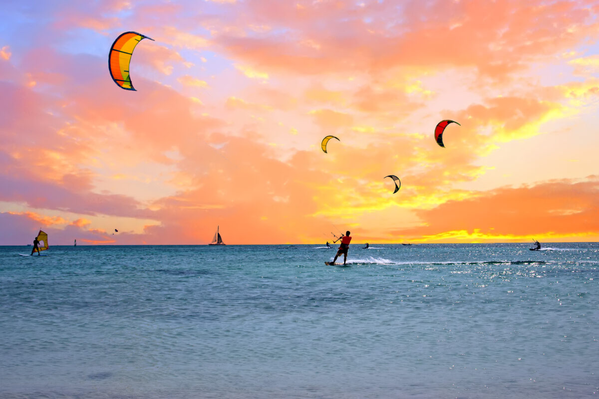 Kiteboarders riding the waves in Aruba at sunset with colorful kites flying above the ocean and an orange-pink sky in the background.