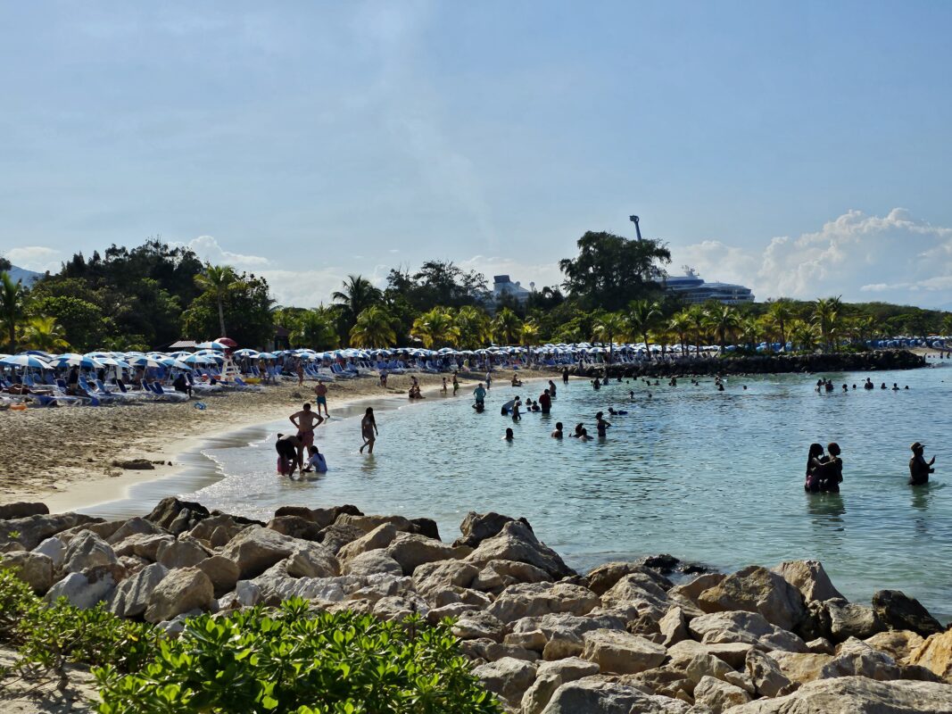Shoreline scene at Labadee featuring palm trees, rocky foreground, and cruise guests enjoying a beach day near docked ships in the distance.