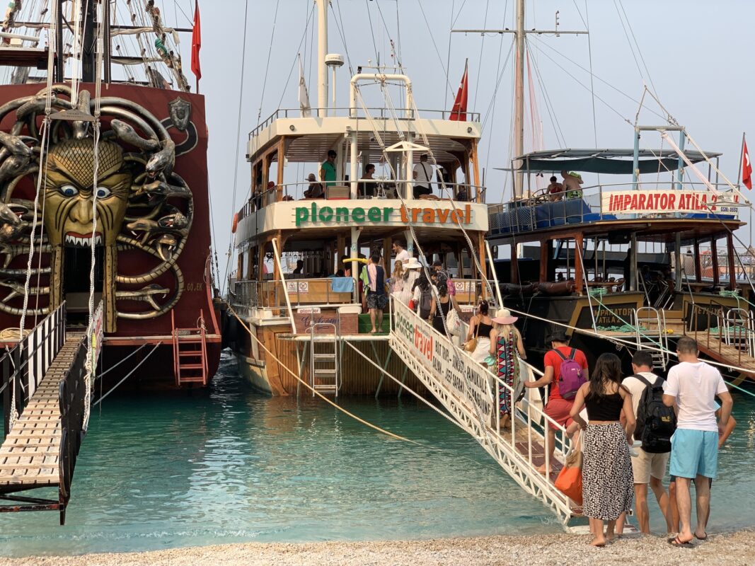 Tourists boarding wooden excursion boats via gangplanks at a small dock, with one boat featuring a large, painted monster face.