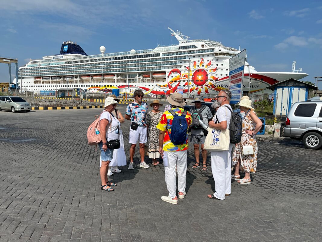 Small group of passengers in sun hats and cruise wear standing in circle with tour guide, waiting at the port beside a colorful cruise ship with a painted sun design.