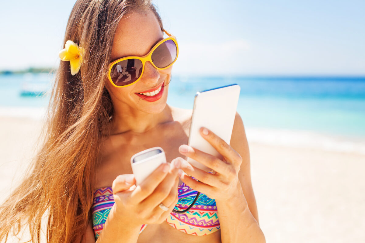 Beachgoer with sunglasses and long hair using a portable phone charger, demonstrating one of the key beach essentials for staying connected and prepared.