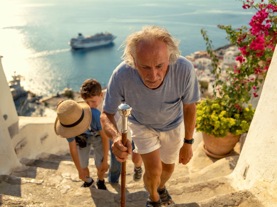 Older man with a cane climbing steep steps with two kids and a cruise ship below, illustrating cruise excursion mistakes for multigenerational families.