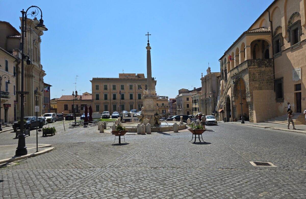 A wide cobblestone piazza in Tarquinia with a tall Baroque fountain topped by an obelisk in the center, framed by historic buildings.