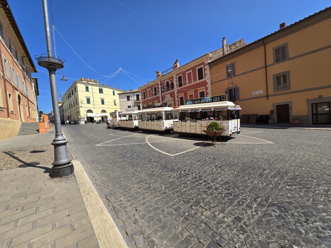 A white tourist trolley train drives through a cobblestone square in Tarquinia, Italy, surrounded by pastel-colored buildings in yellow, peach, and red, under a clear blue sky.
