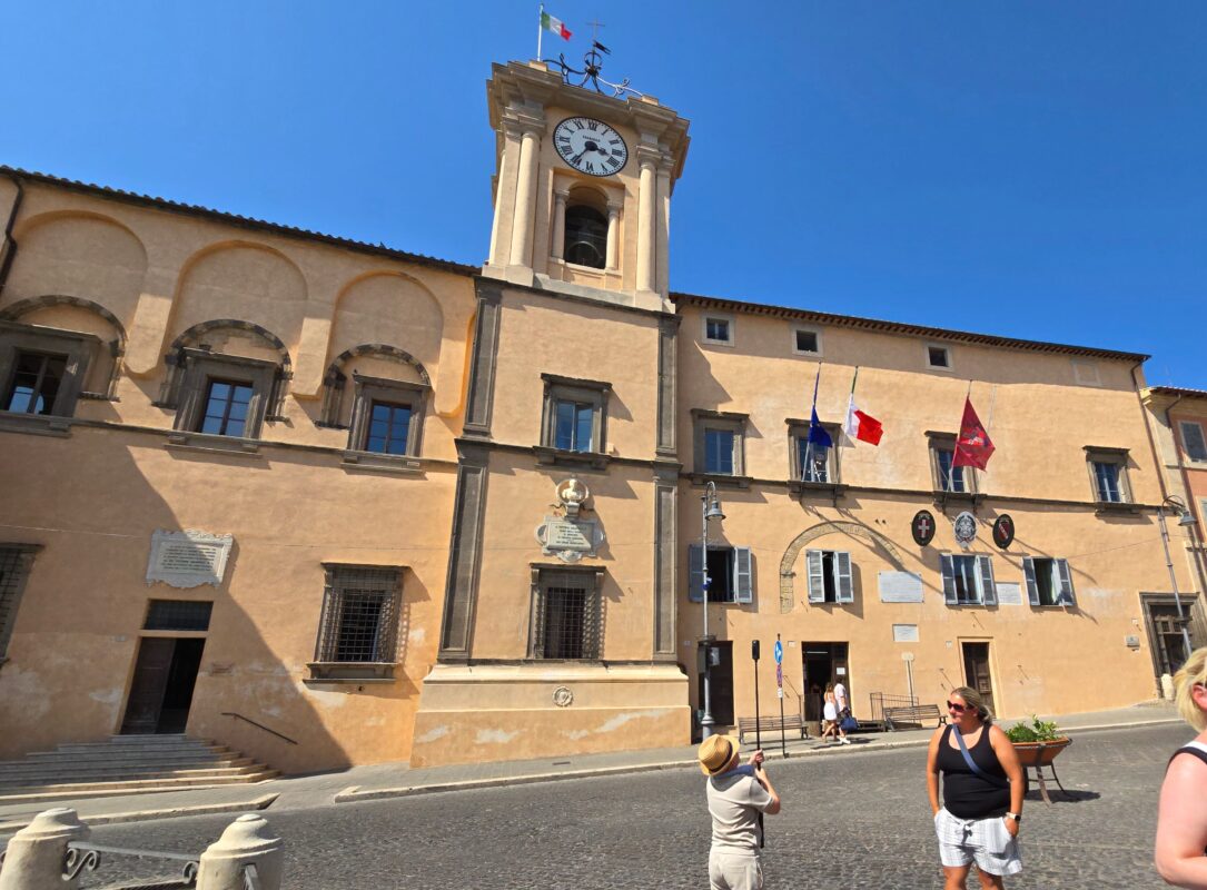 A sunlit square in Tarquinia with the Town Hall’s ochre façade, arched windows, and tall clock tower flying the Italian flag.