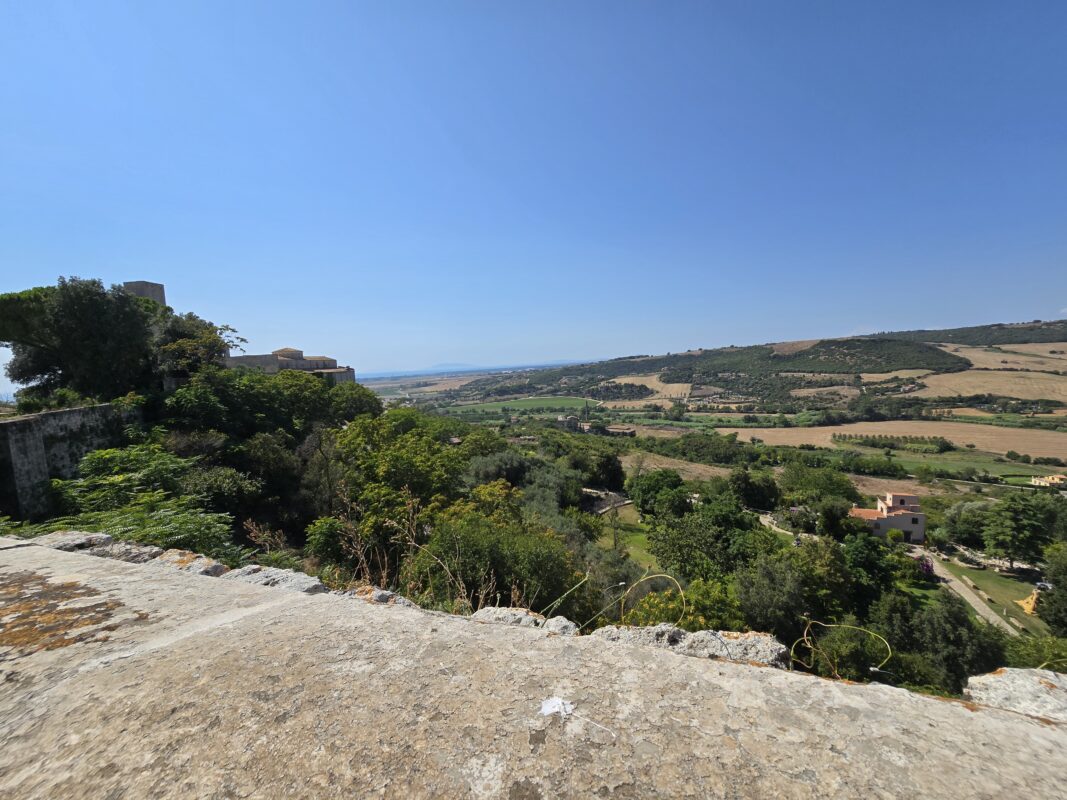 Overlook from Tarquinia’s medieval walls onto a patchwork of green and brown fields, clusters of trees, and farmhouses, with the horizon fading into the sea.