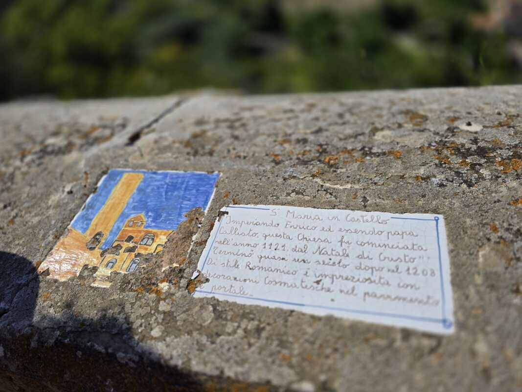 Close-up of a small hand-painted tile and handwritten note on a stone wall, depicting Santa Maria in Castello church with its tall Romanesque tower.