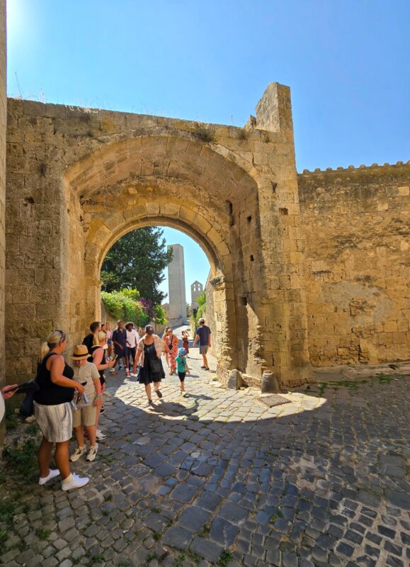 A towering rectangular medieval stone tower in Tarquinia, rising above a sunlit piazza marked with circular cobblestone patterns.