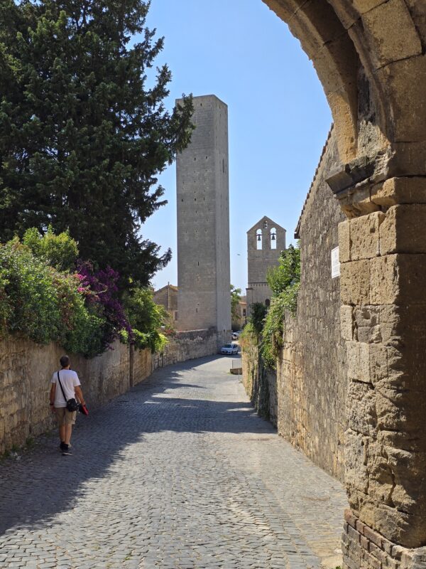 View along a cobblestone lane in Tarquinia leading to a tall medieval stone tower and church bell gable, with a man walking under an archway.