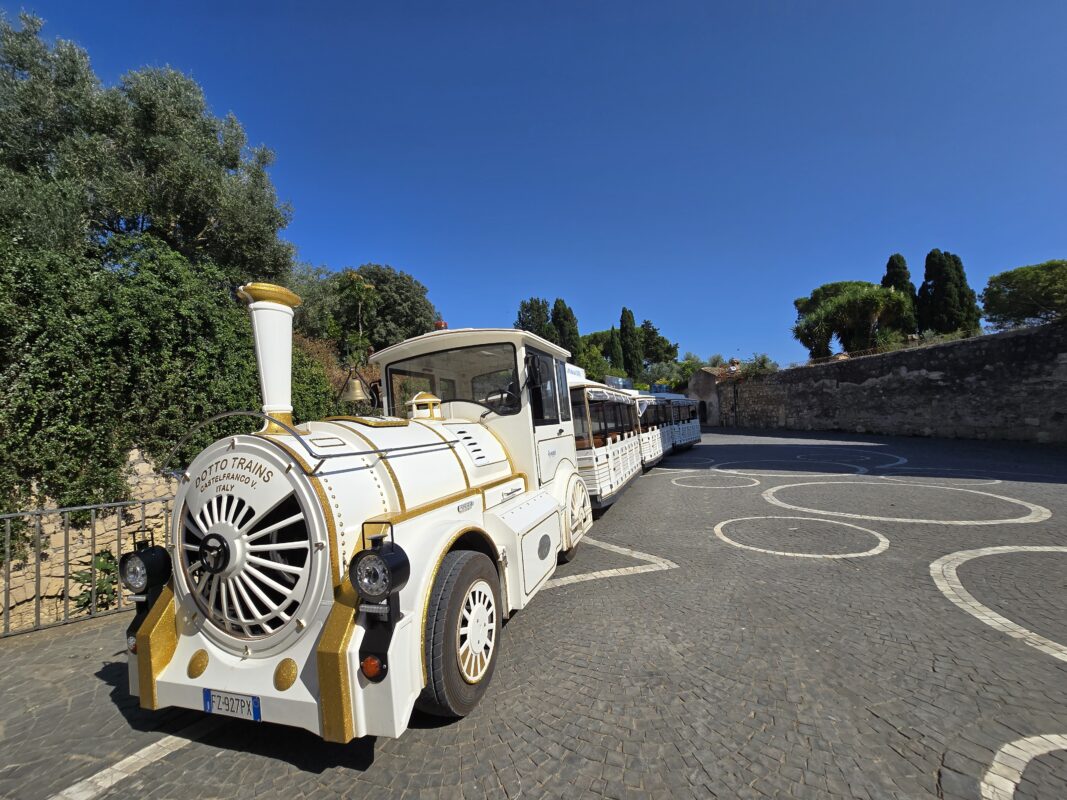 A white-and-gold tourist trolley train parked on a cobblestone square in Tarquinia, with medieval stone walls and greenery in the background.