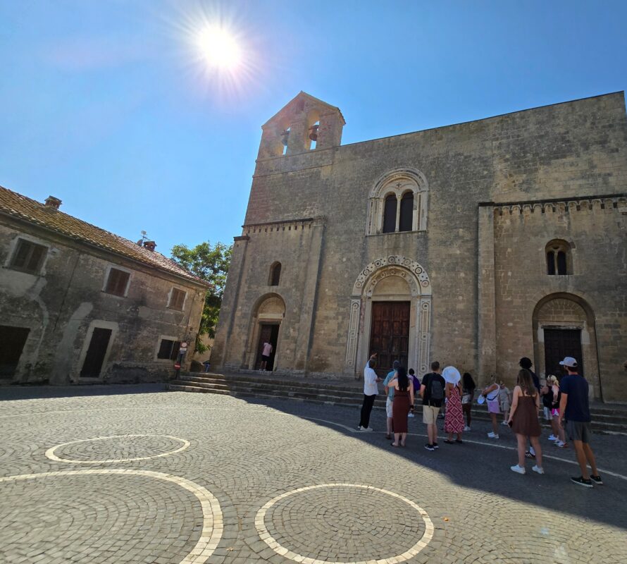 A group of cruise visitors stands in front of Santa Maria in Castello, a 12th-century stone church in Tarquinia with arched doorways, a simple bell tower, and decorative carvings around the main entrance under a bright sun.