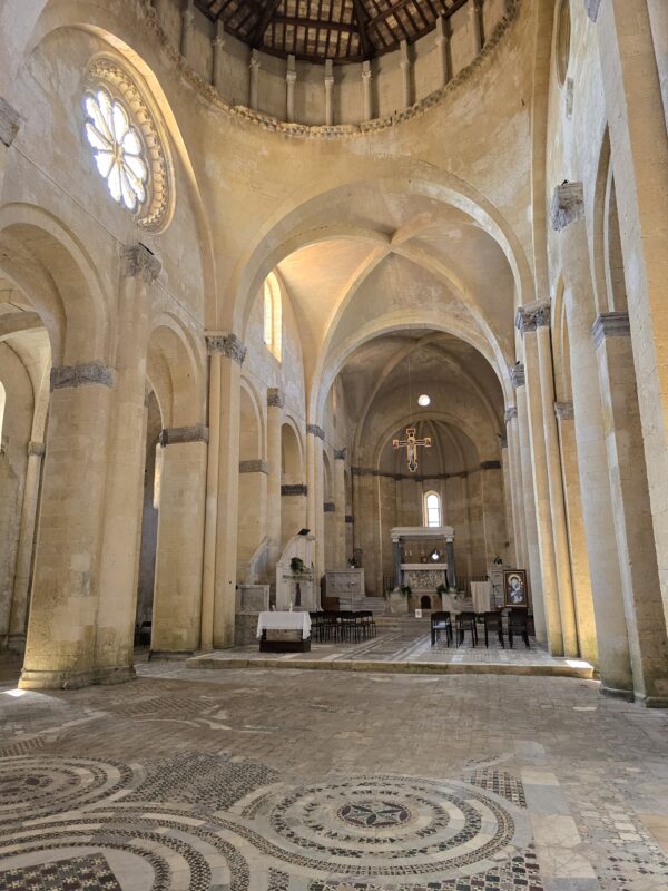 Interior of Santa Maria in Castello in Tarquinia, showing tall Romanesque arches, a large rose window casting light into the nave, and the main altar in the distance beneath a vaulted ceiling.