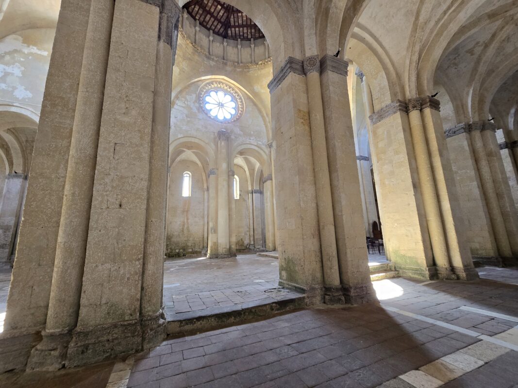 Side aisle of Santa Maria in Castello in Tarquinia, with a glowing rose window above, thick stone pillars, and shafts of light illuminating the Romanesque architecture.