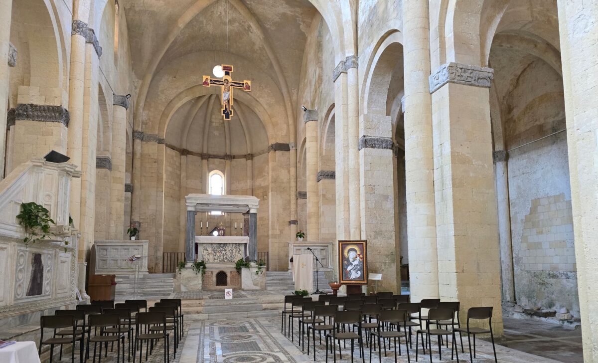 The altar of Santa Maria in Castello, featuring a marble structure, a suspended crucifix above, rows of chairs for worshippers, and detailed mosaic floors leading up to the sanctuary.