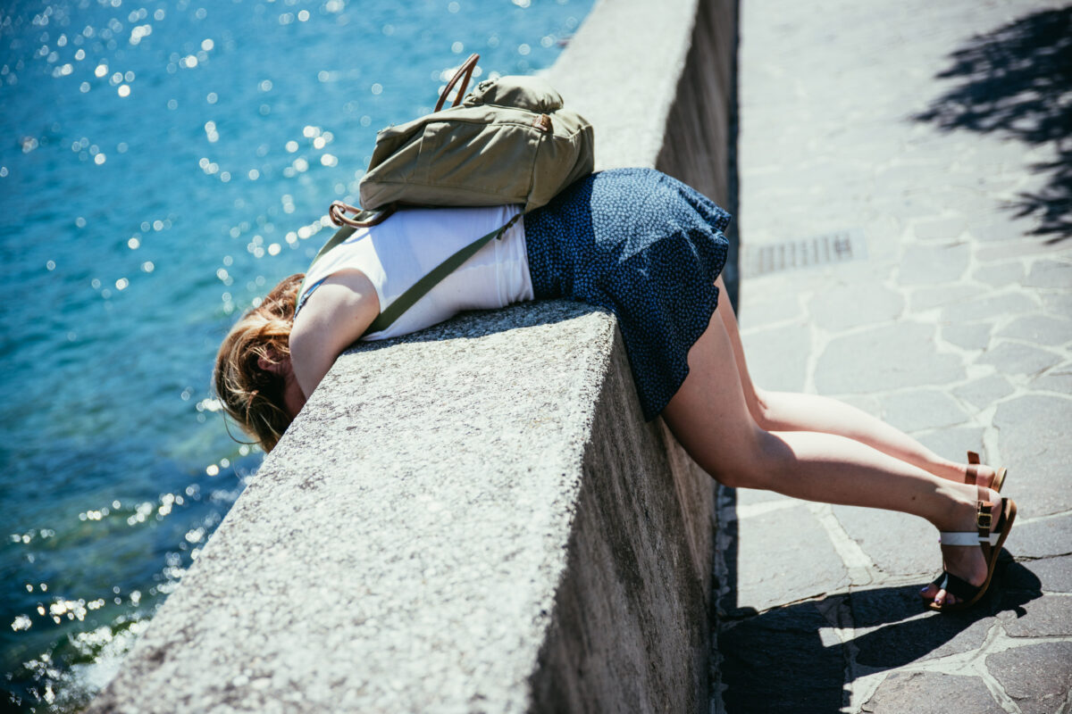 Woman leaning over a seaside promenade wall with backpack, showing fatigue from a long day of sightseeing.