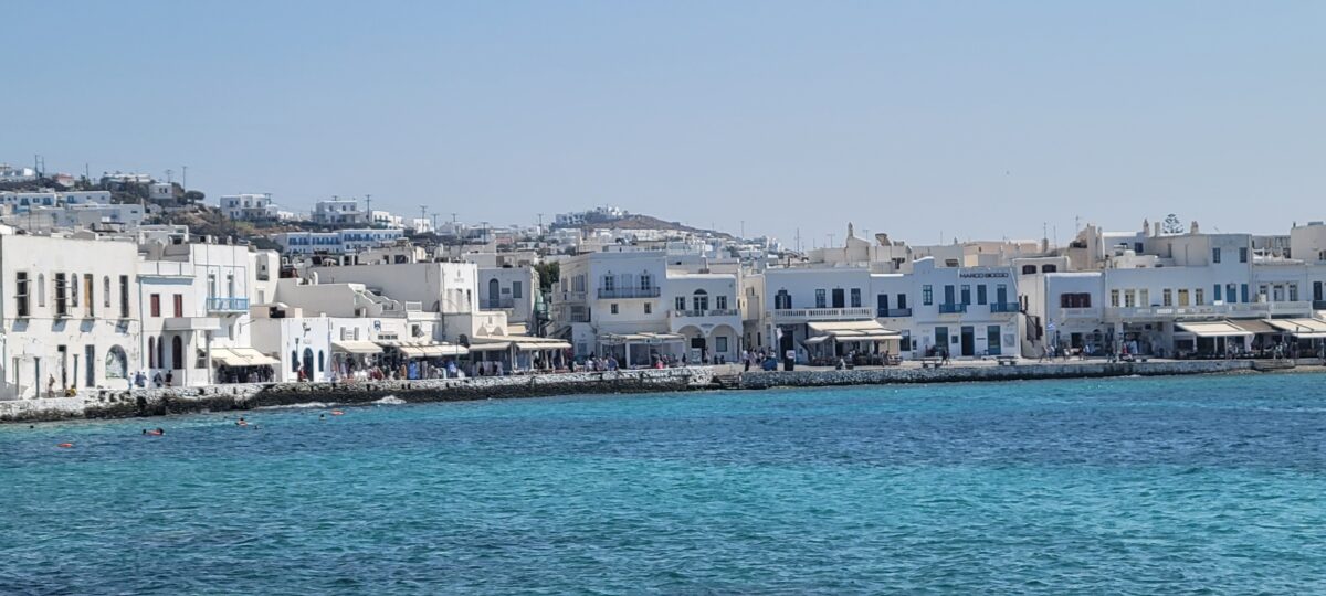 Whitewashed buildings on the coast of Mykonos, Greece, with turquoise water in the foreground, a highlight of Mediterranean cruise excursions.
