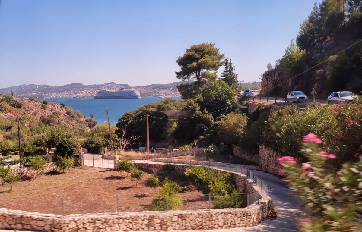 View of Cephalonia coastline with cruise ship anchored offshore and winding hillside road with cars.