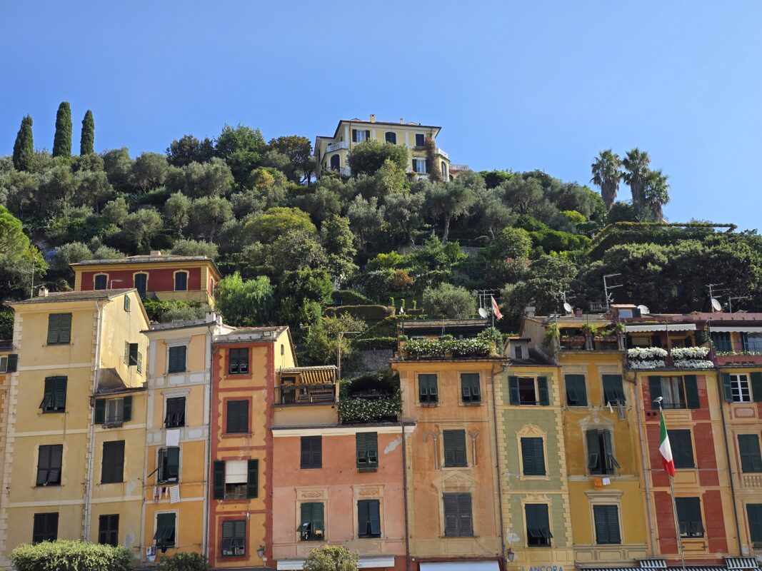 A dramatic view of the Portofino cruise port, with colorful pastel waterfront homes tightly clustered along the harbor. Behind them, steep hillsides rise sharply, layered with terraced gardens, winding stone paths, and lush greenery, topped by elegant villas overlooking the Italian Riviera.