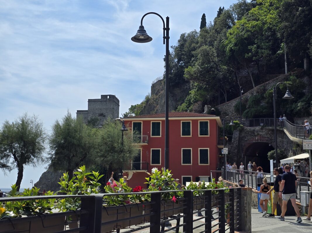 View of a red house, flowers, and castle ruins above Monterosso with people walking nearby.