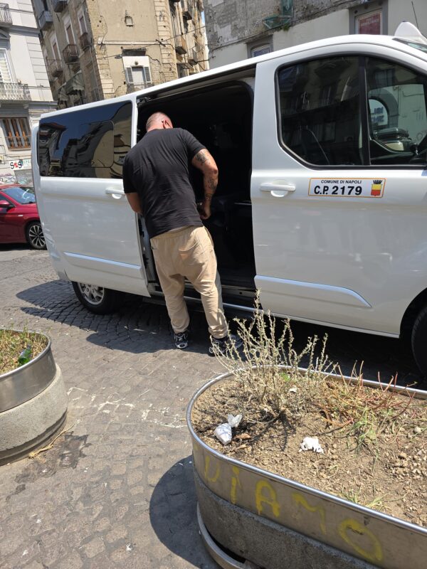 A man in a black shirt and tan pants stands beside a white taxi van on a cobblestone street in Naples, preparing to close the side door.