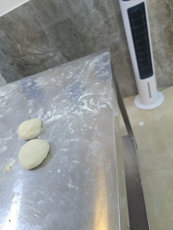 Two small balls of raw pizza dough resting on a floured stainless steel countertop in a cooking classroom.