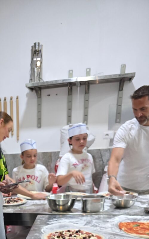 Children in chef hats and aprons standing at a stainless steel counter, adding toppings to their pizzas during a cooking class in Naples.
